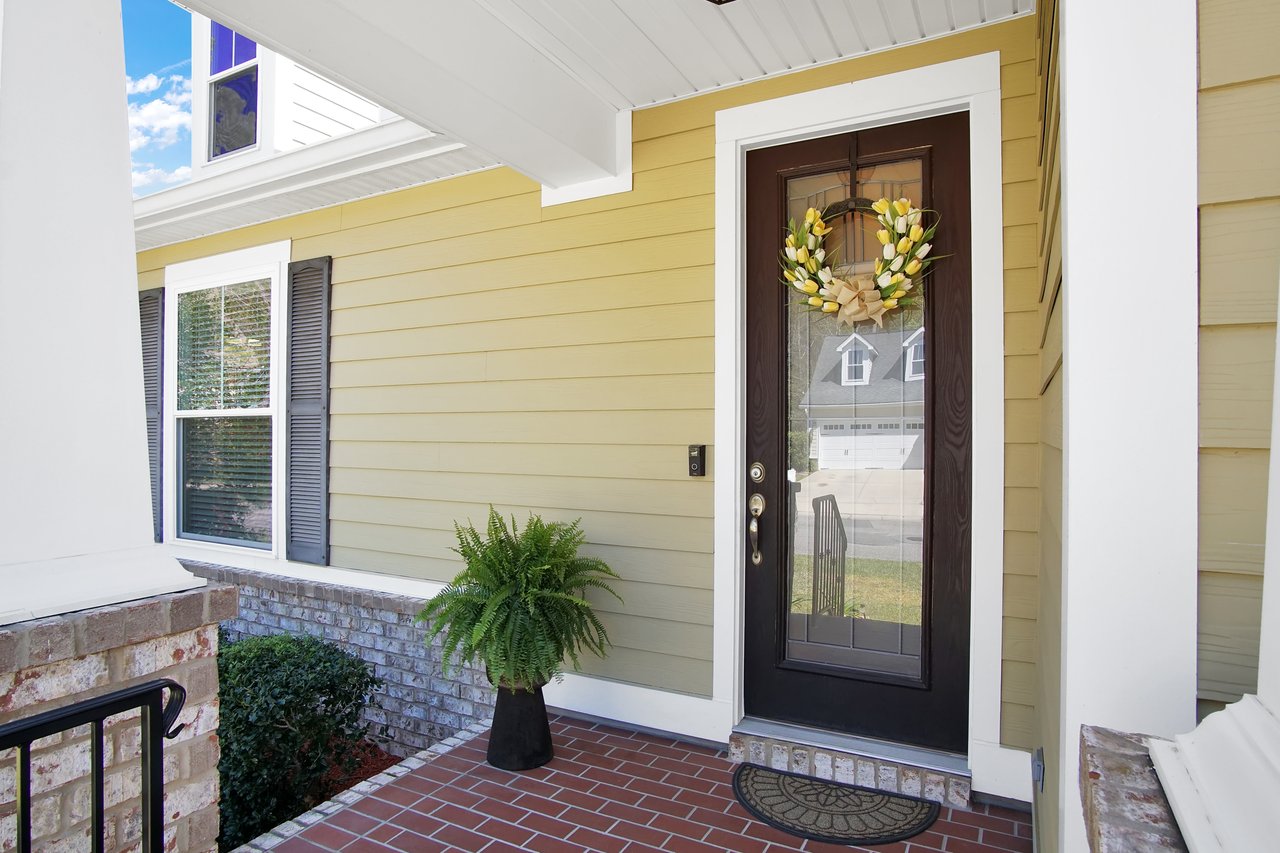 Front porch with a glass door featuring a yellow floral wreath. Adjacent to the door is a leafy green potted fern. The setting is welcoming and tidy.