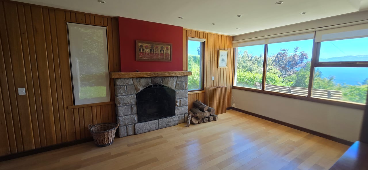 Living room with a stone fireplace, wood-paneled walls, and broad windows overlooking lake and mountains.