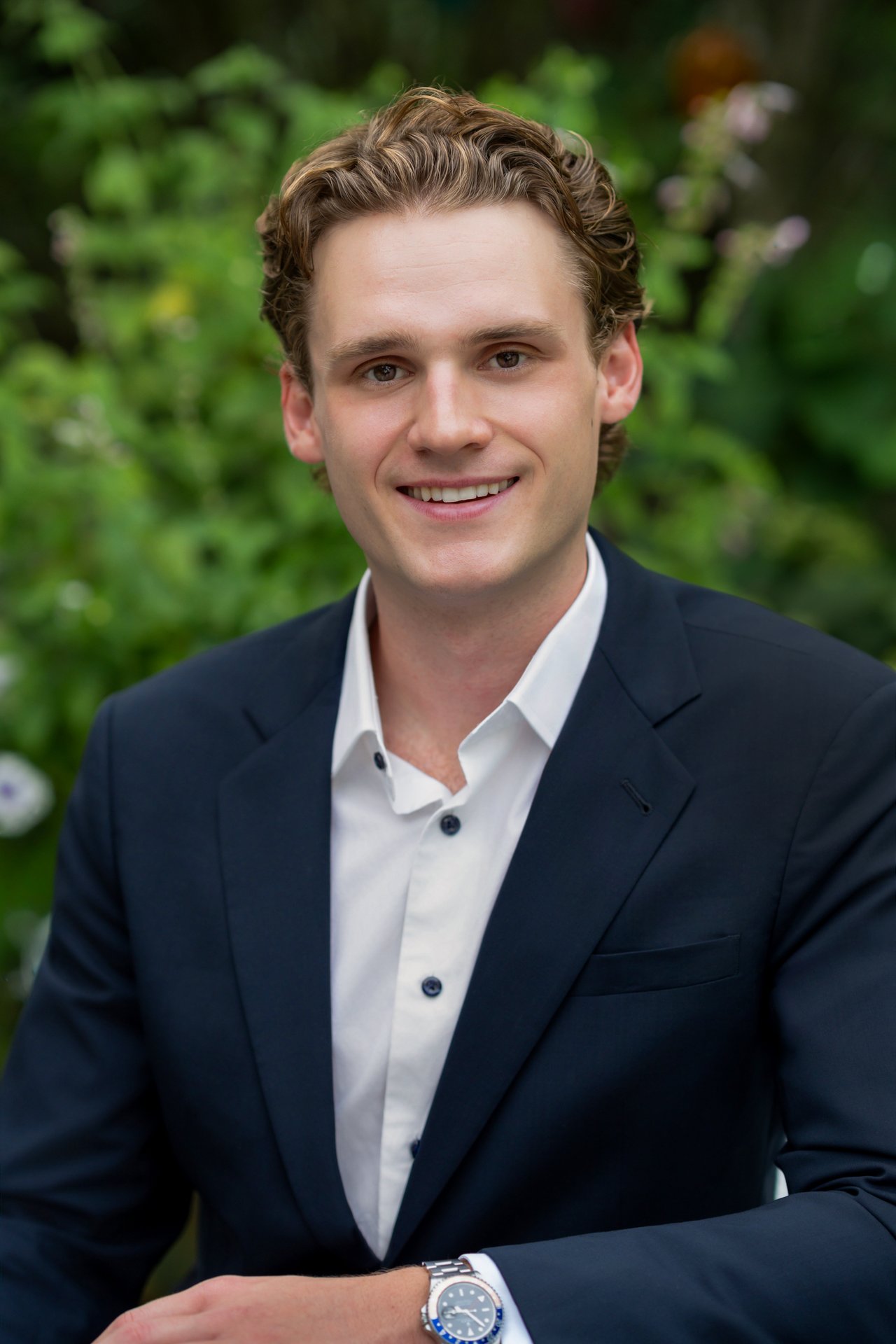 Gavin Feller in a navy suit and white shirt, smiling against leafy greenery.