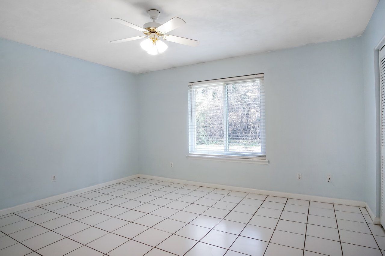 An empty room with light blue walls and white tiled floor. A ceiling fan with lights is on, and a window with blinds overlooks greenery outside.