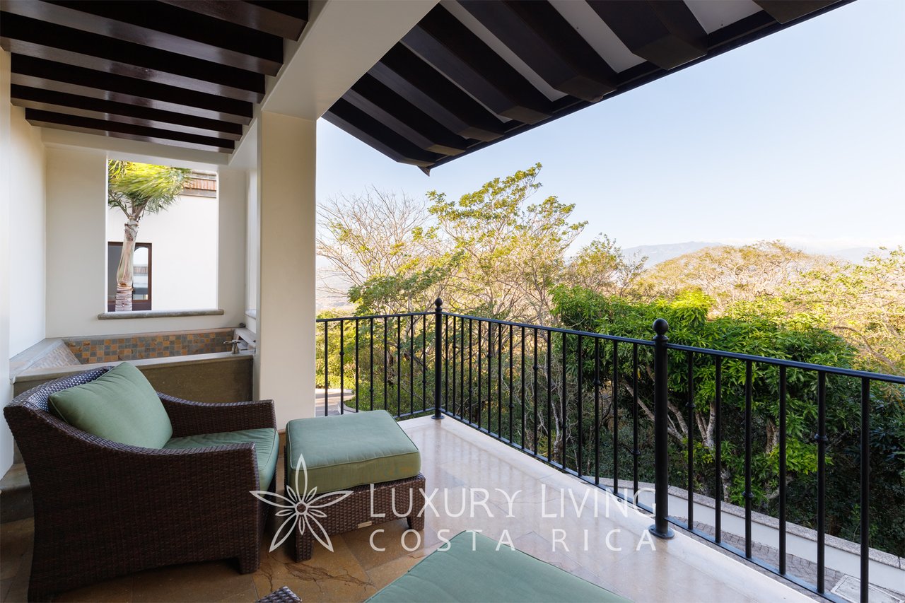 Covered terrace with wicker seating and a view over treetops toward distant hills.