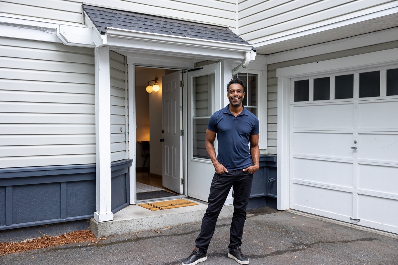 White-sided house with an open front door and attached garage; a man stands in the driveway.