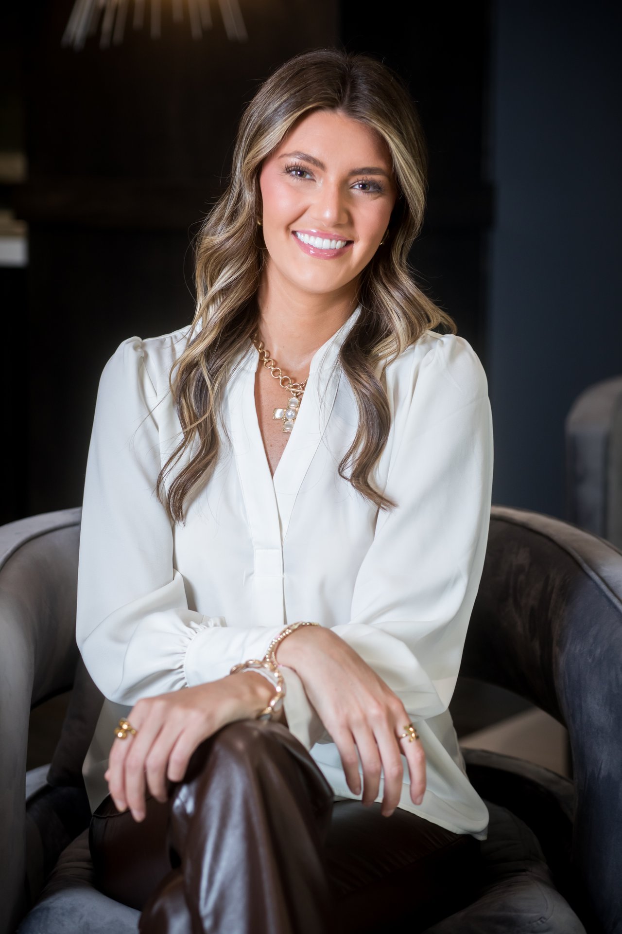 Katie Ullman seated in a dark chair, wearing a white blouse and gold jewelry.