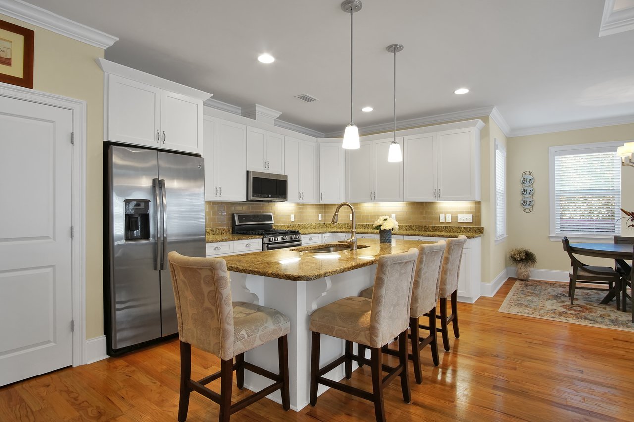 Spacious kitchen with white cabinets and stainless steel appliances. An island with granite countertop and pendant lights, beige bar stools, and a dining area.