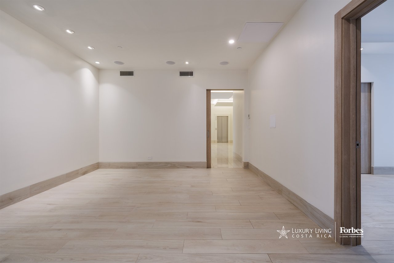 Empty apartment room with pale wood-look flooring, recessed lights, and doorways to adjoining spaces.