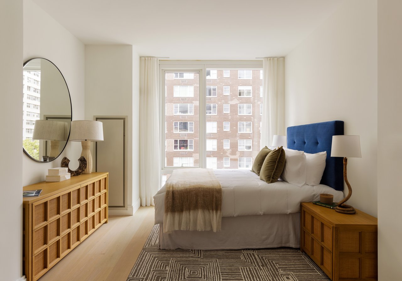 Staged bedroom with a blue headboard, warm wood furniture, and windows facing a brick apartment building.