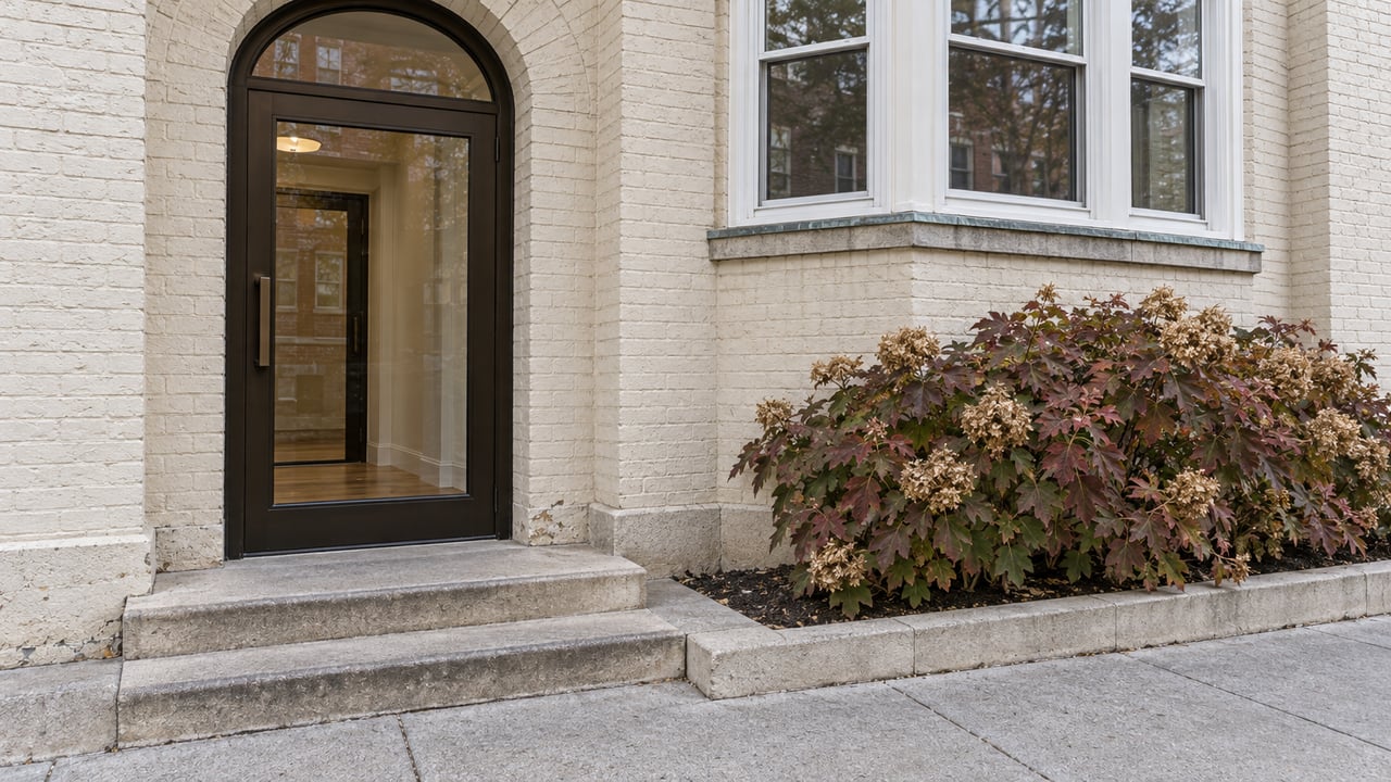 Off-white brick apartment entrance with an arched glass door, limestone steps, projecting bay windows, and an oakleaf hydrangea beside the sidewalk.