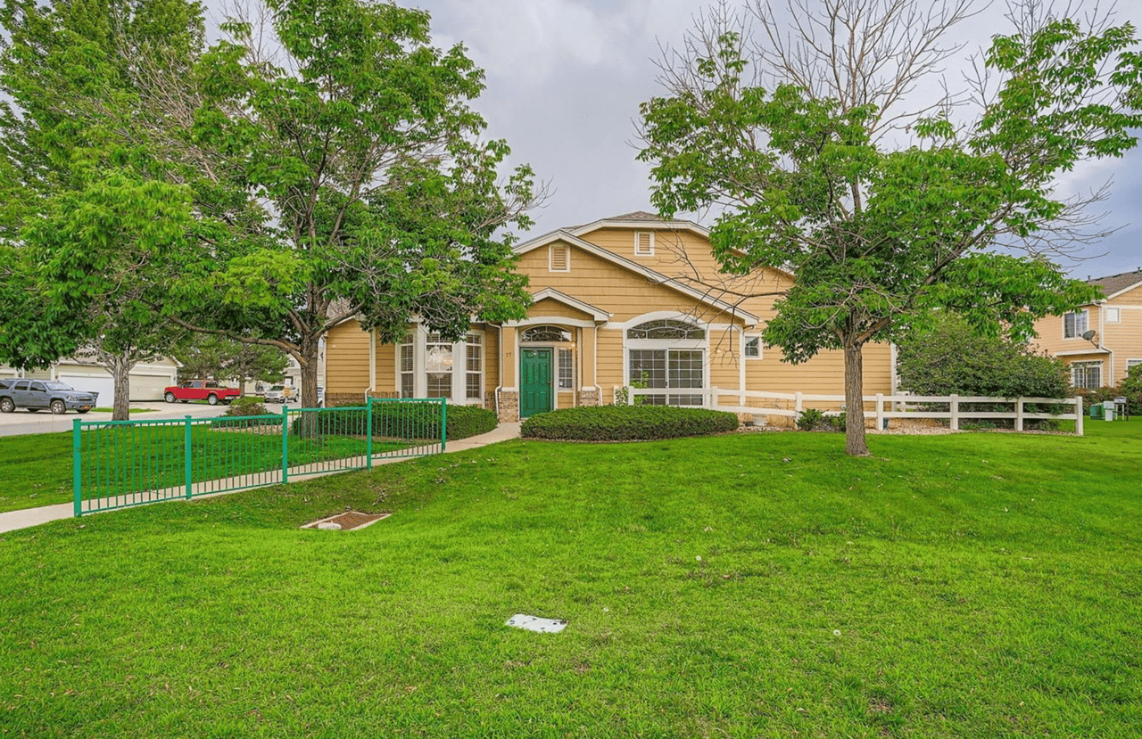 Yellow two-story residential building with a green door, framed by mature trees and a broad lawn.