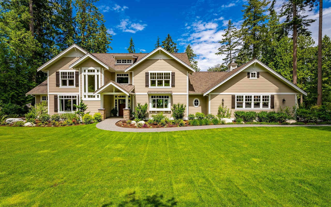 Beige two-story home with multiple gables, landscaped garden beds, and a broad green lawn.