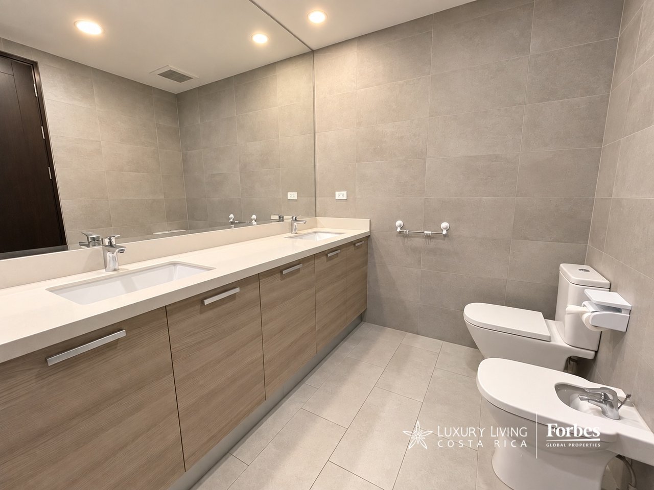 Modern tiled bathroom with a double-sink wood vanity, toilet, and bidet.