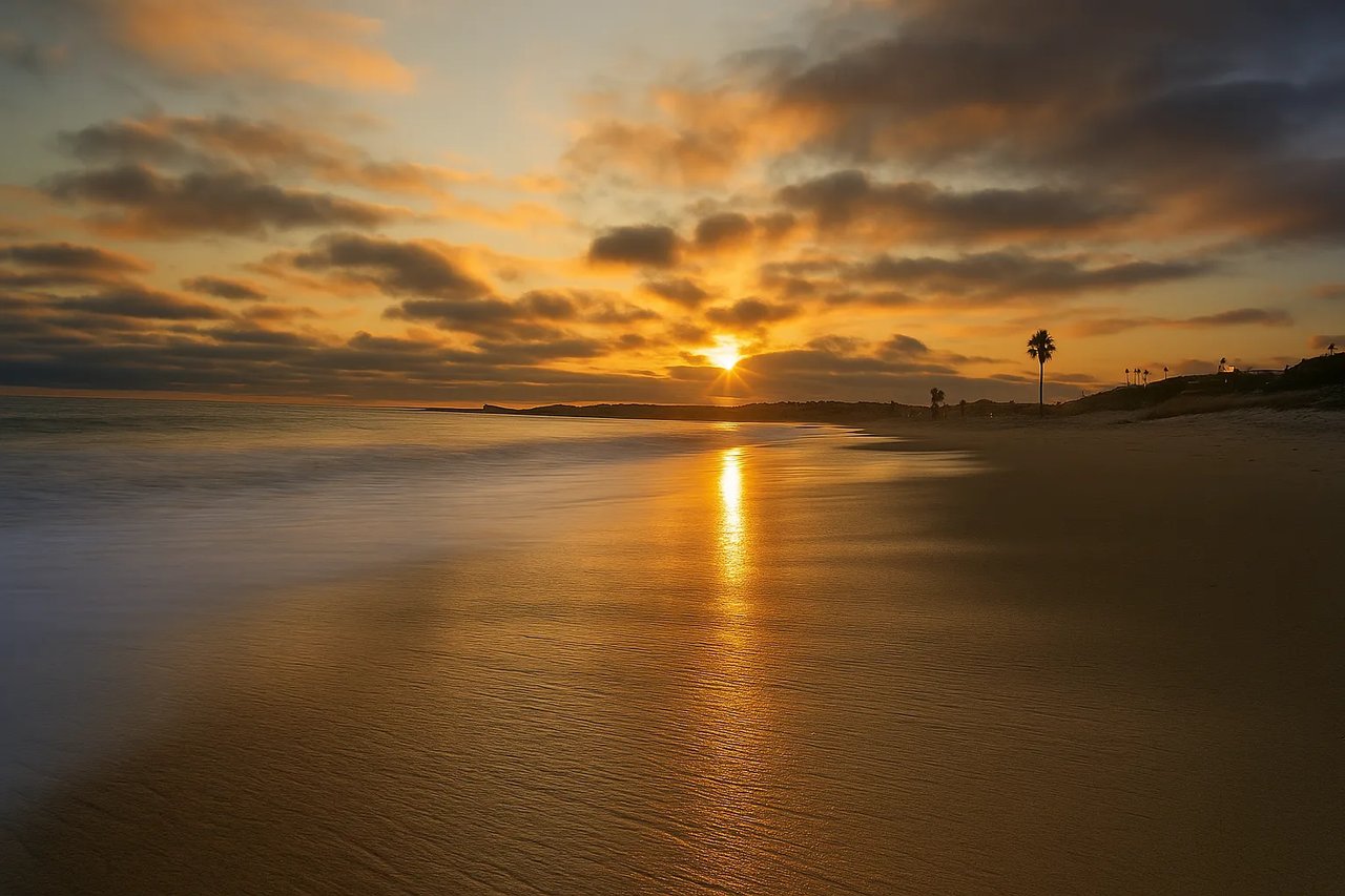 Sunset over Capistrano Beach Park in Dana Point, California, with golden light reflecting on the wet sand and waves gently rolling ashore.