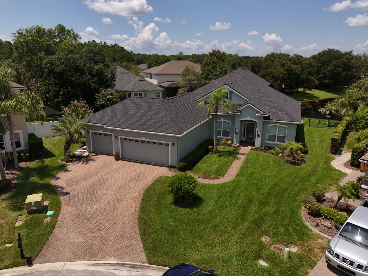 Aerial view of a gray single-story home with a three-car garage, brick-paver driveway, and landscaped lawn.