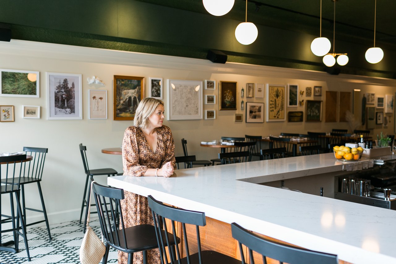 Cait Berry of Insiders Realty sitting thoughtfully at a clean, modern bar counter with her hands clasped, looking contemplatively into a styled cafe space featuring a gallery wall of botanical art. Her composed and strategic posture represents the careful planning and advisory role she plays in helping Verona homeowners navigate the complex sequence of buying and selling a home at the same time.