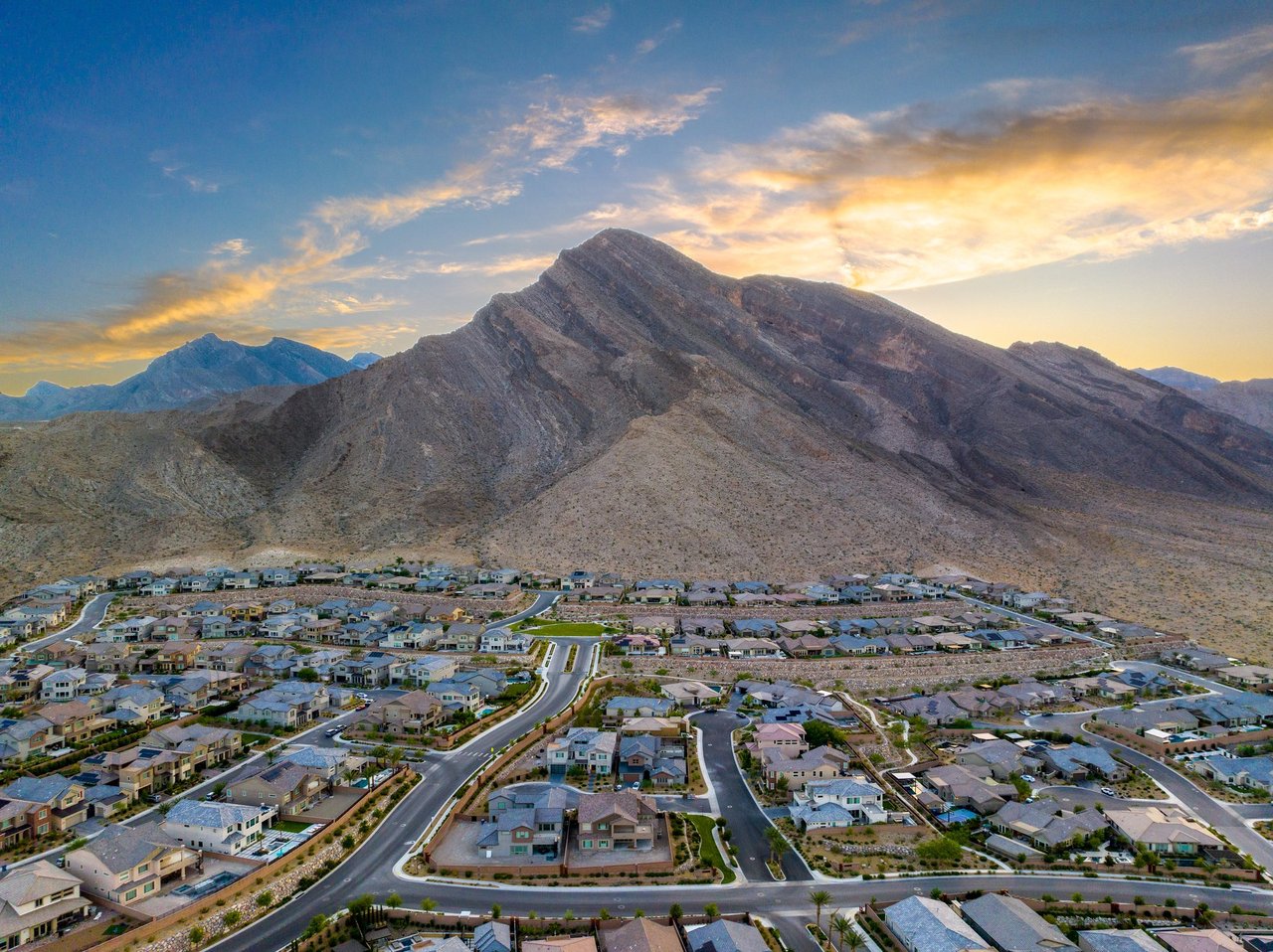 Aerial view of Reverence homes in Summerlin at sunset, with curving streets, a green park and a rugged desert mountain rising directly behind the neighborhood on the northwest edge of Las Vegas
