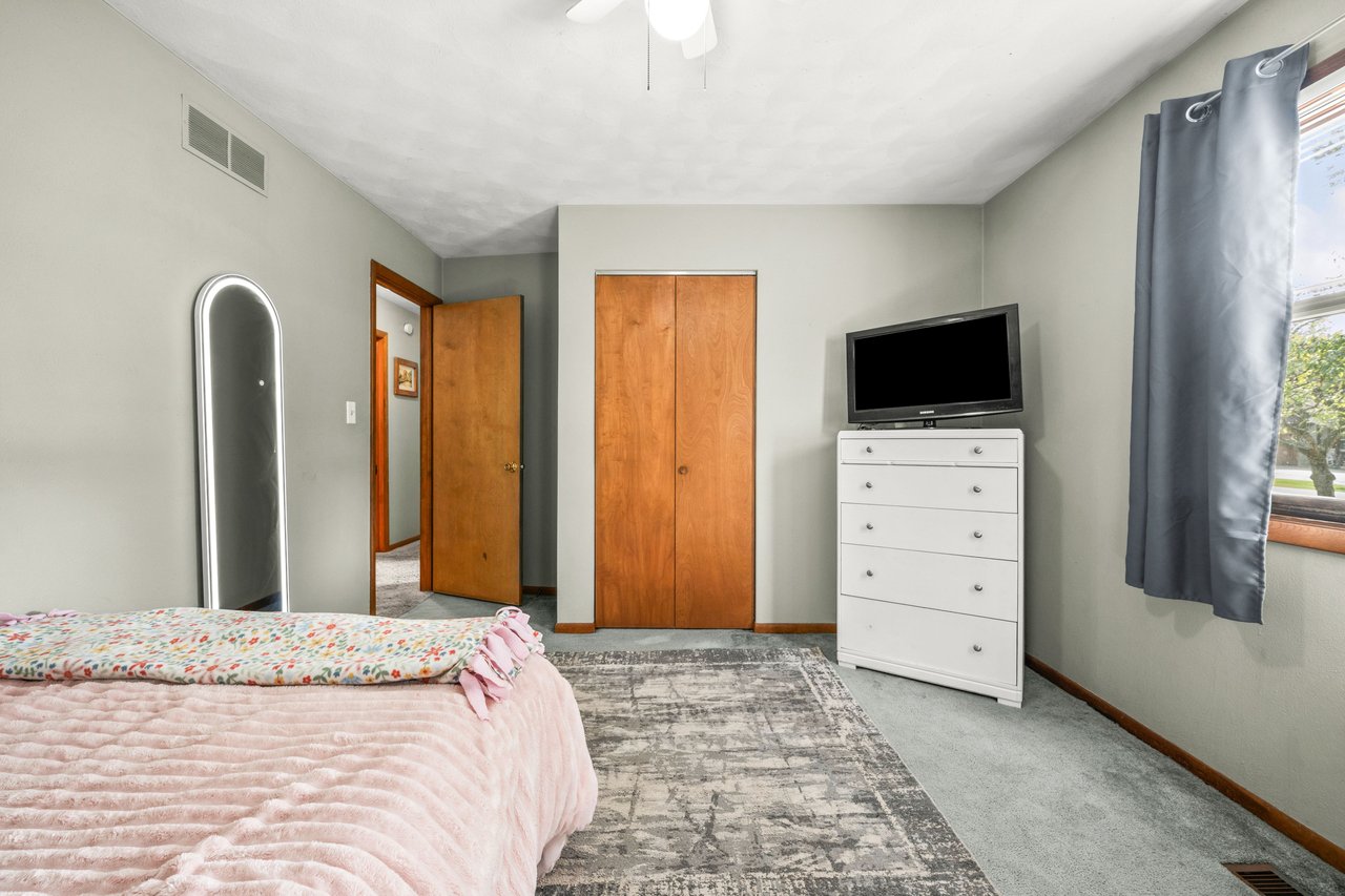 Bedroom with a pink quilted bed, wooden closet doors, and a white dresser with a television.