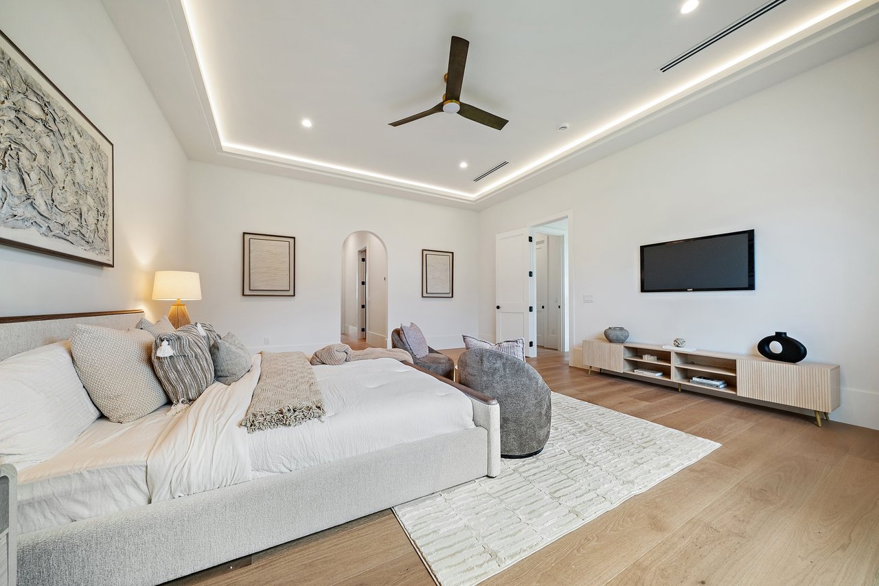 Staged bedroom with a large upholstered bed, tray ceiling, ceiling fan, and wall-mounted television.