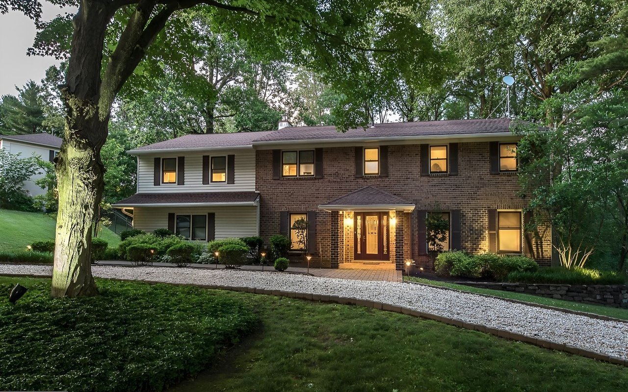 Two-story brick home with illuminated windows and a curved gravel walkway beneath mature trees.