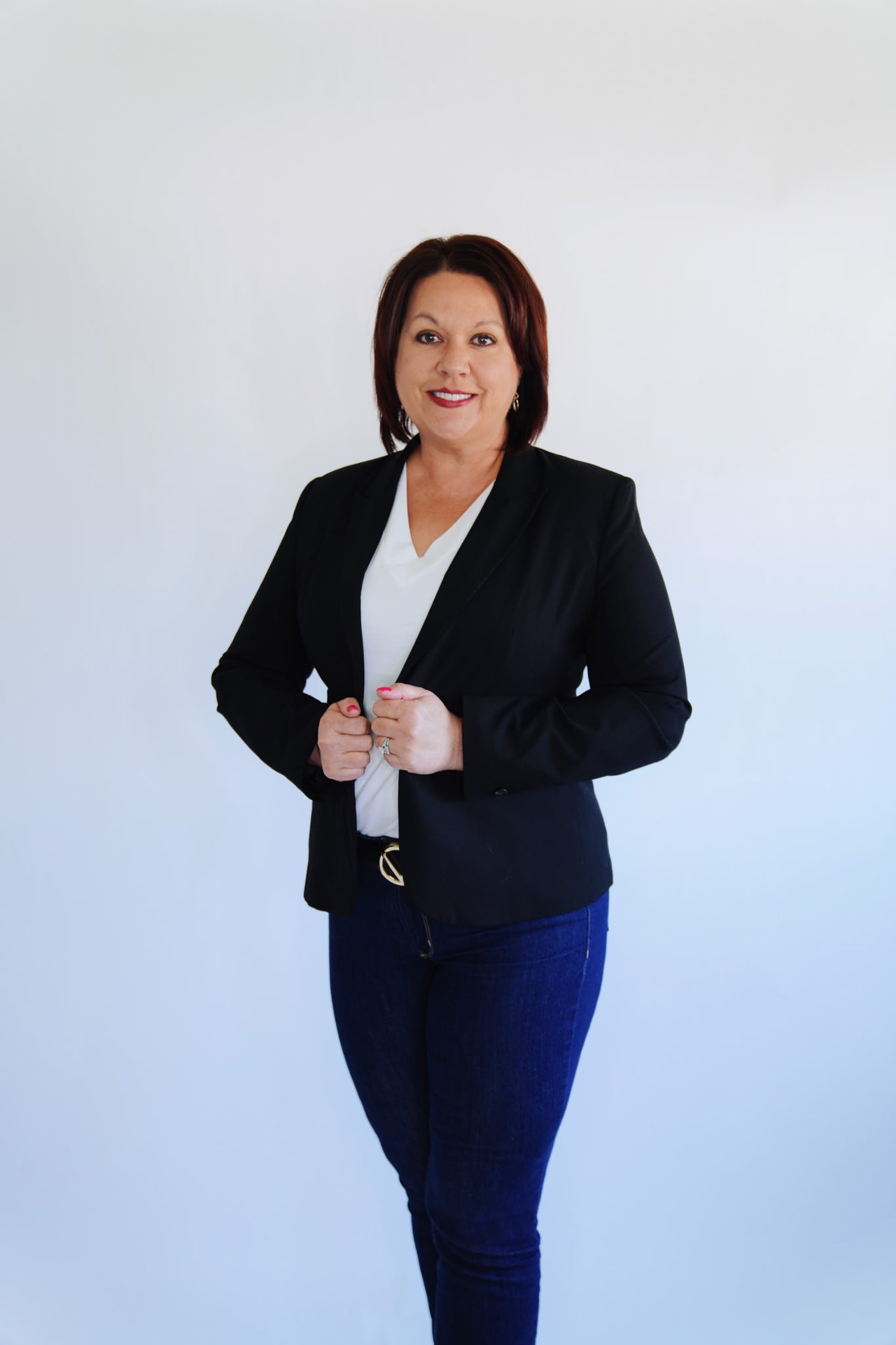 Kristen Flavin in a black blazer and white top, standing against a pale blue background.