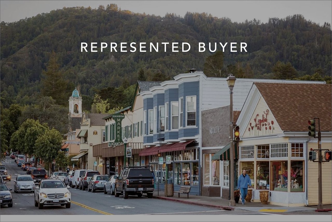 Downtown street lined with storefronts and cars, with wooded hills in the background.