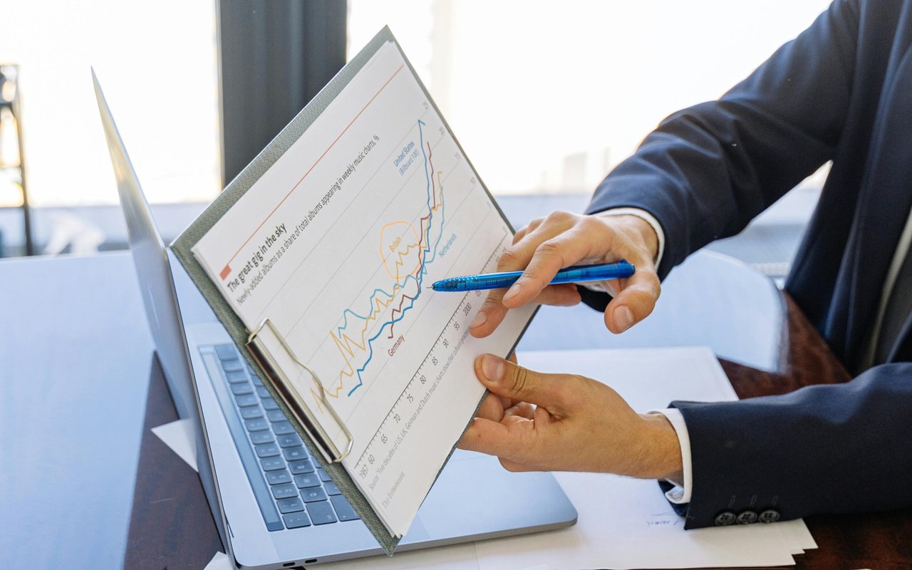 A suited person points to a multicolored financial chart beside an open laptop.