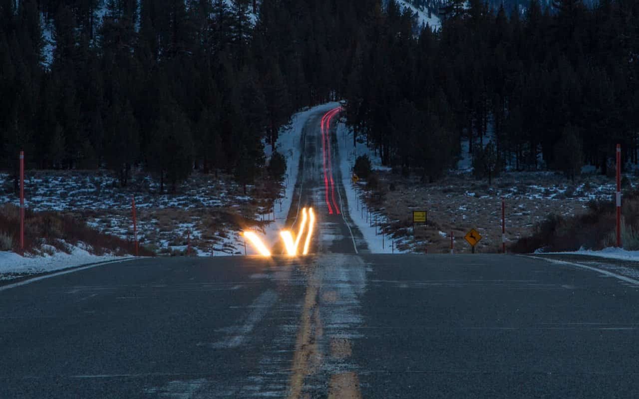 Snow-lined mountain road winding through pine forest at dusk, with red and white light trails.