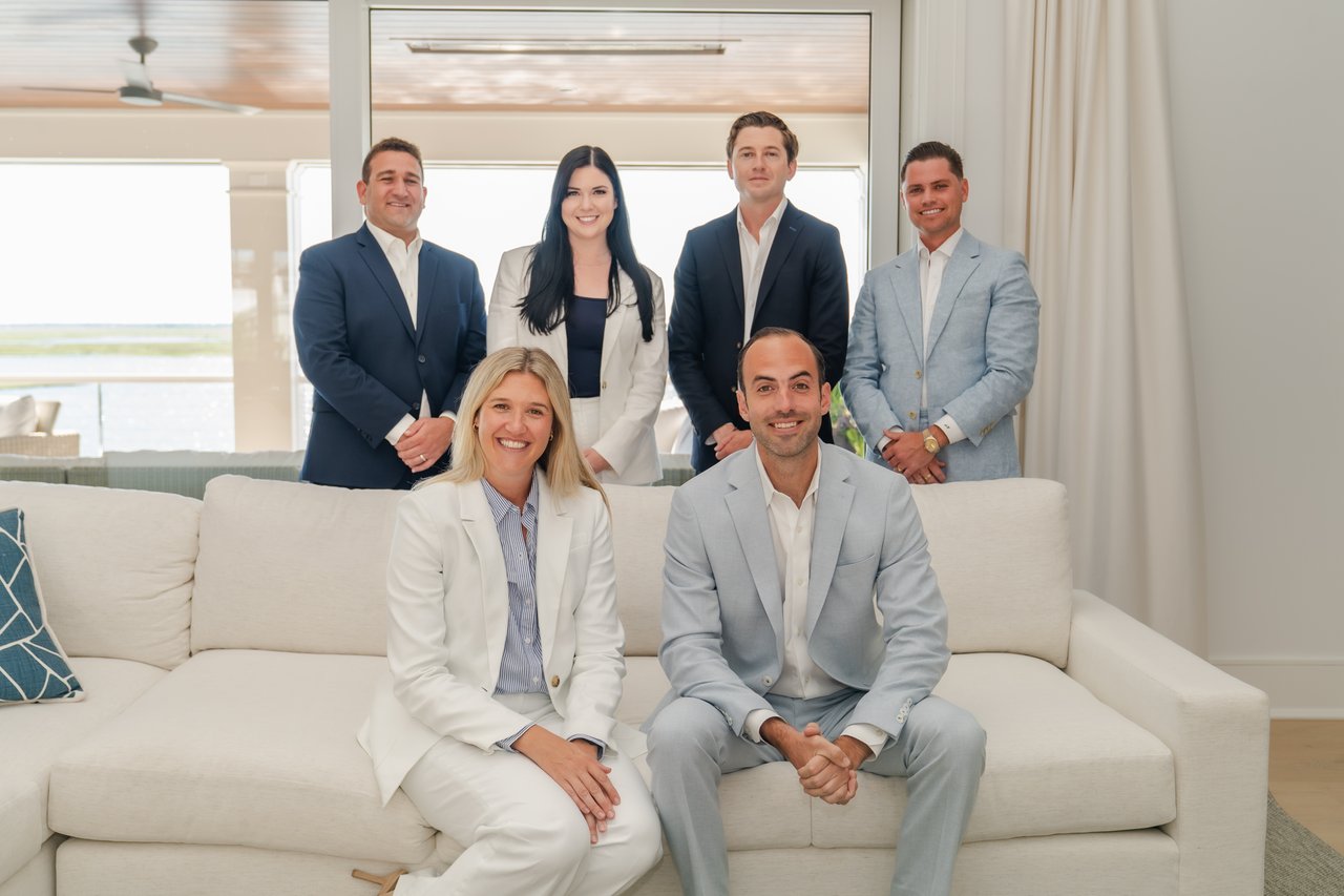 Six real estate professionals pose together around a white sofa in a bright living room.