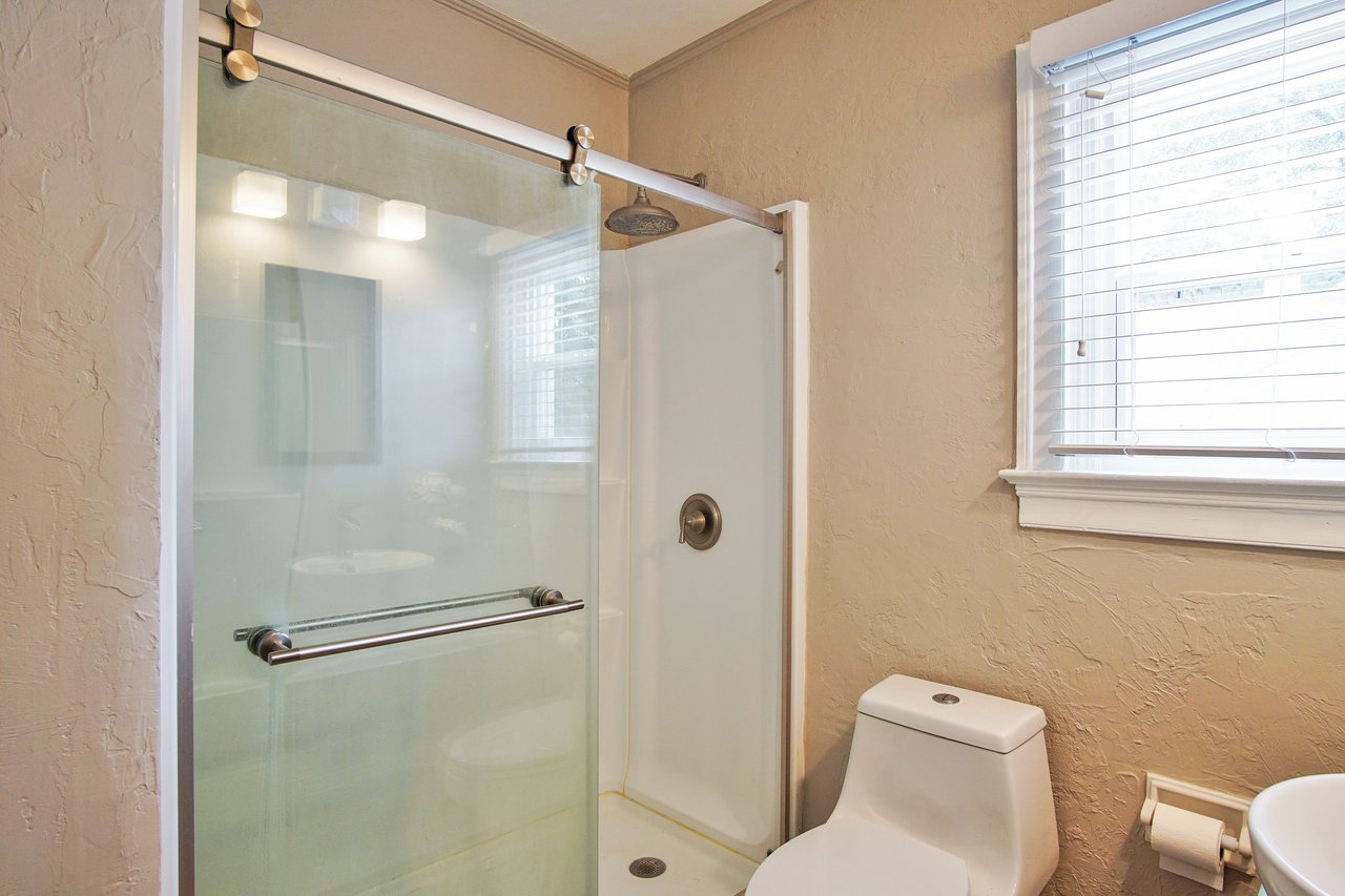 Modern bathroom with a frosted glass shower door, beige textured walls, and a white toilet beside a window with horizontal blinds. Bright and clean.