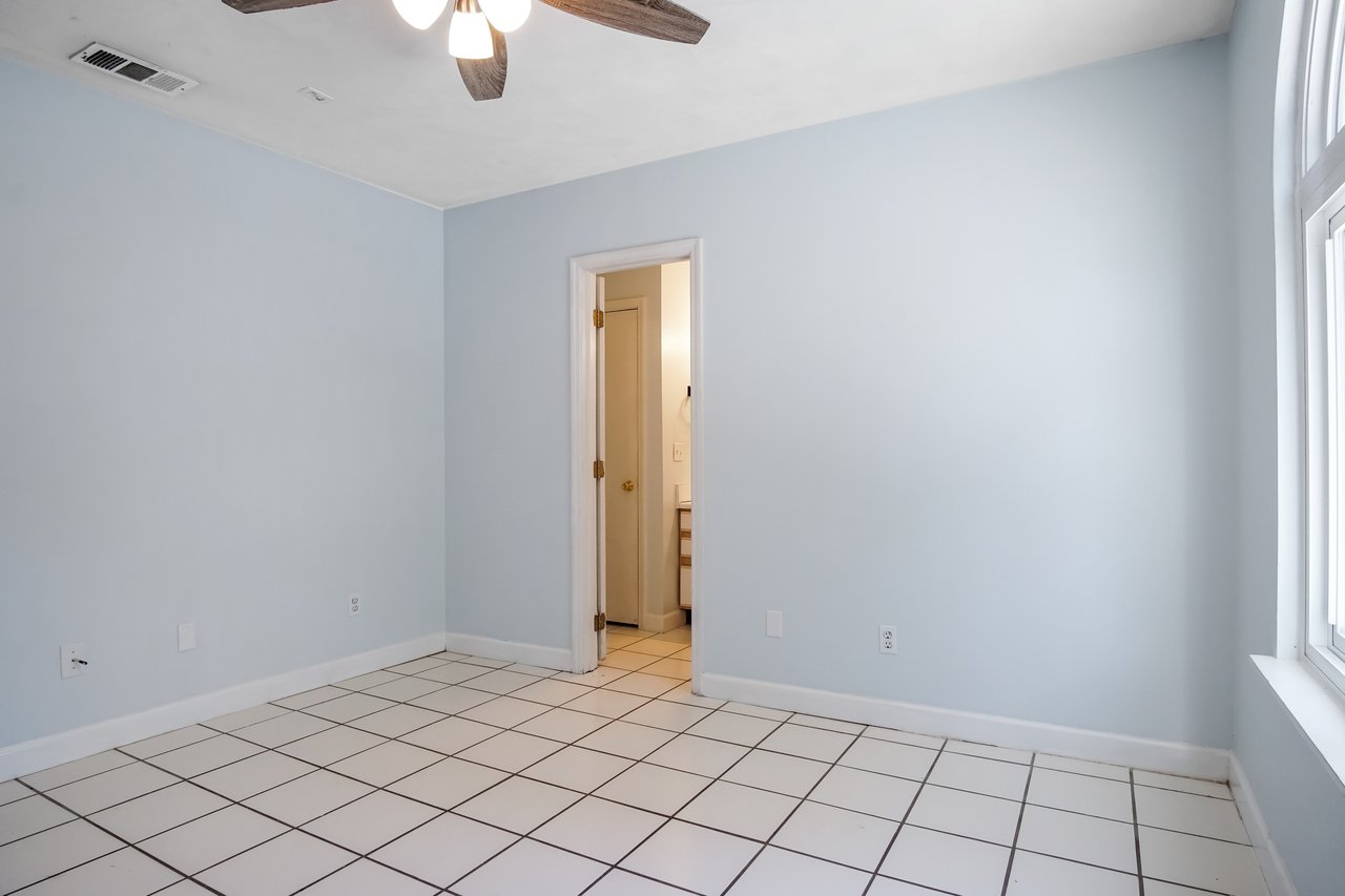 Empty room with light blue walls, white tile floor, and a ceiling fan with lights. An open door leads to a tiled bathroom, creating a clean, minimalist feel.