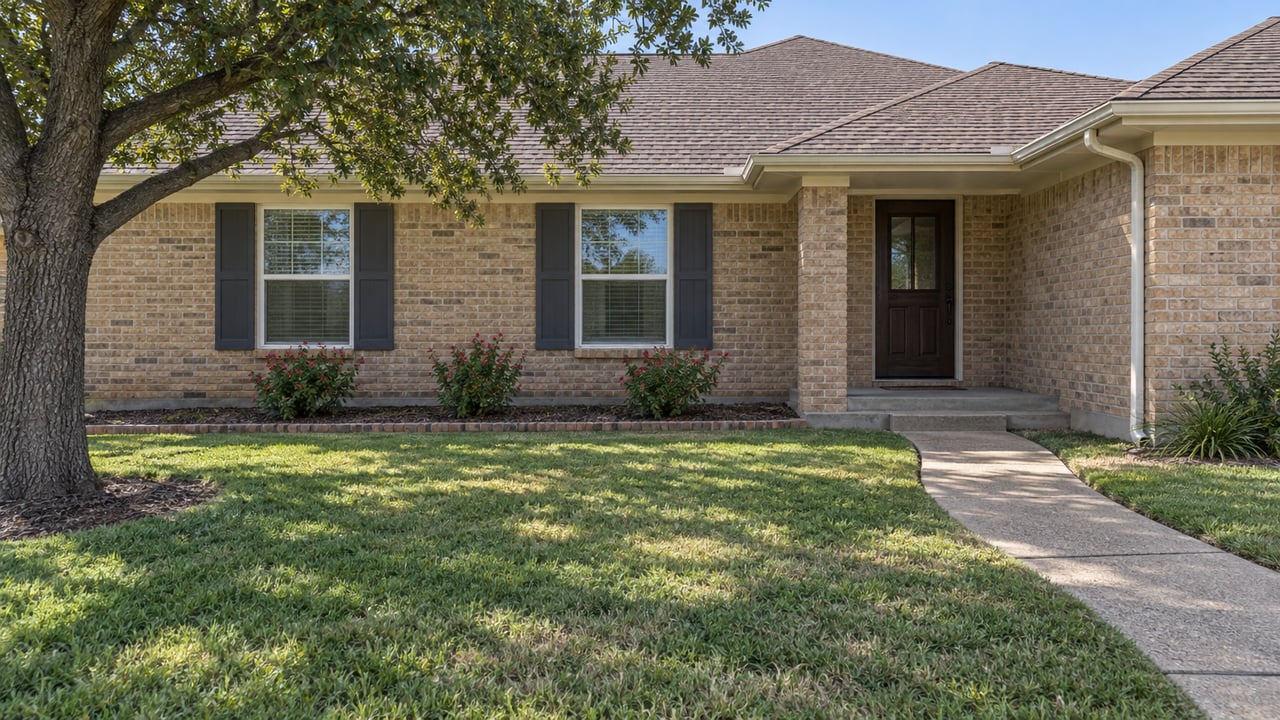 Single-story buff-brick home with a deep front lawn, shaded porch, two shuttered windows and a curving concrete approach.