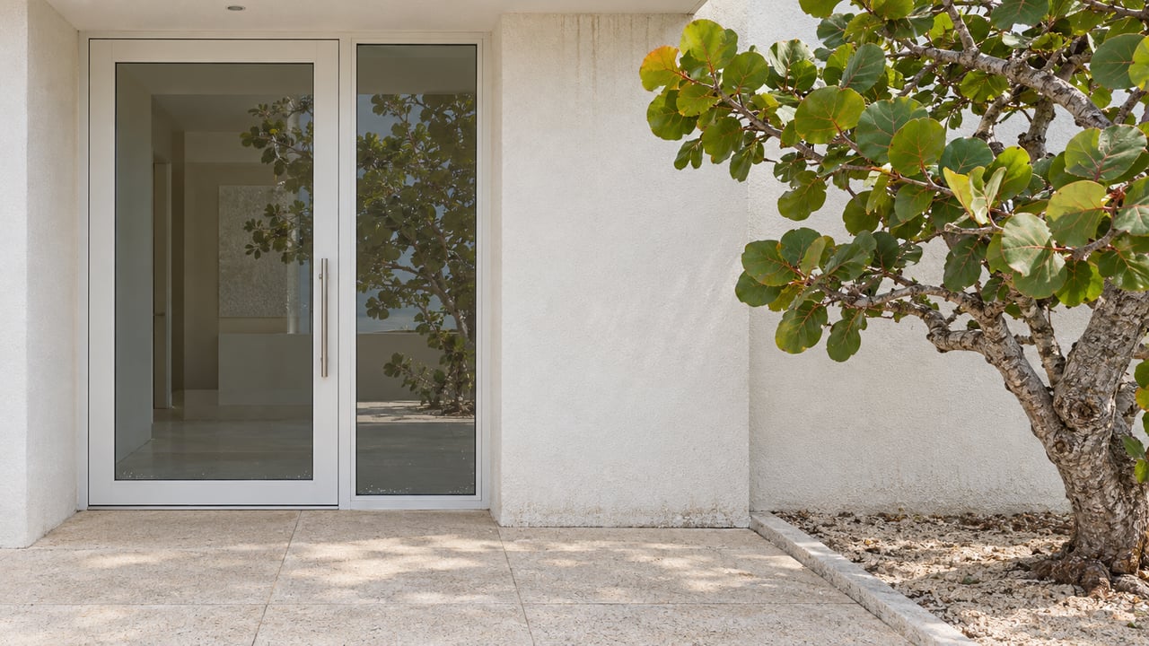 Closed glazed entrance framed by weathered white stucco, pale concrete paving, and one sculptural sea grape tree.