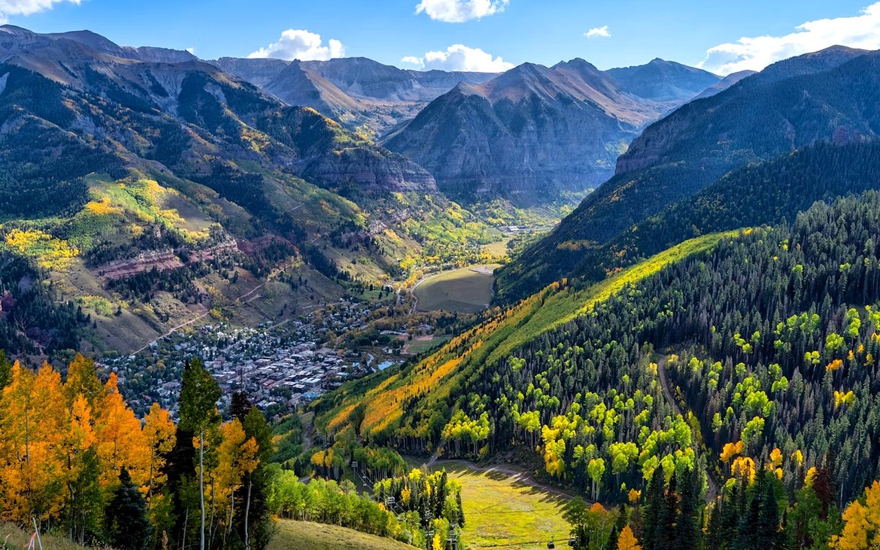 Telluride sits in a mountain valley beneath rugged peaks, with golden autumn trees covering the slopes.
