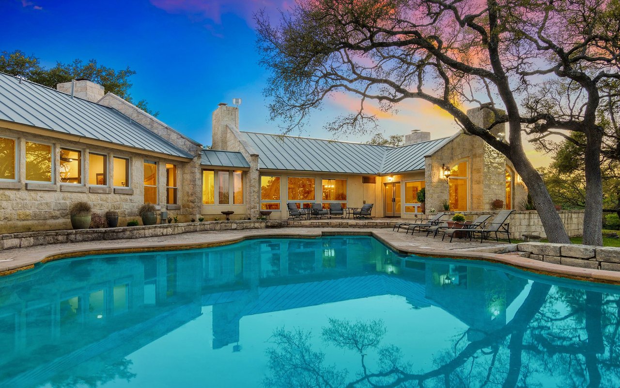 Stone home surrounding a swimming pool, with outdoor lounge chairs and a large oak tree at sunset.