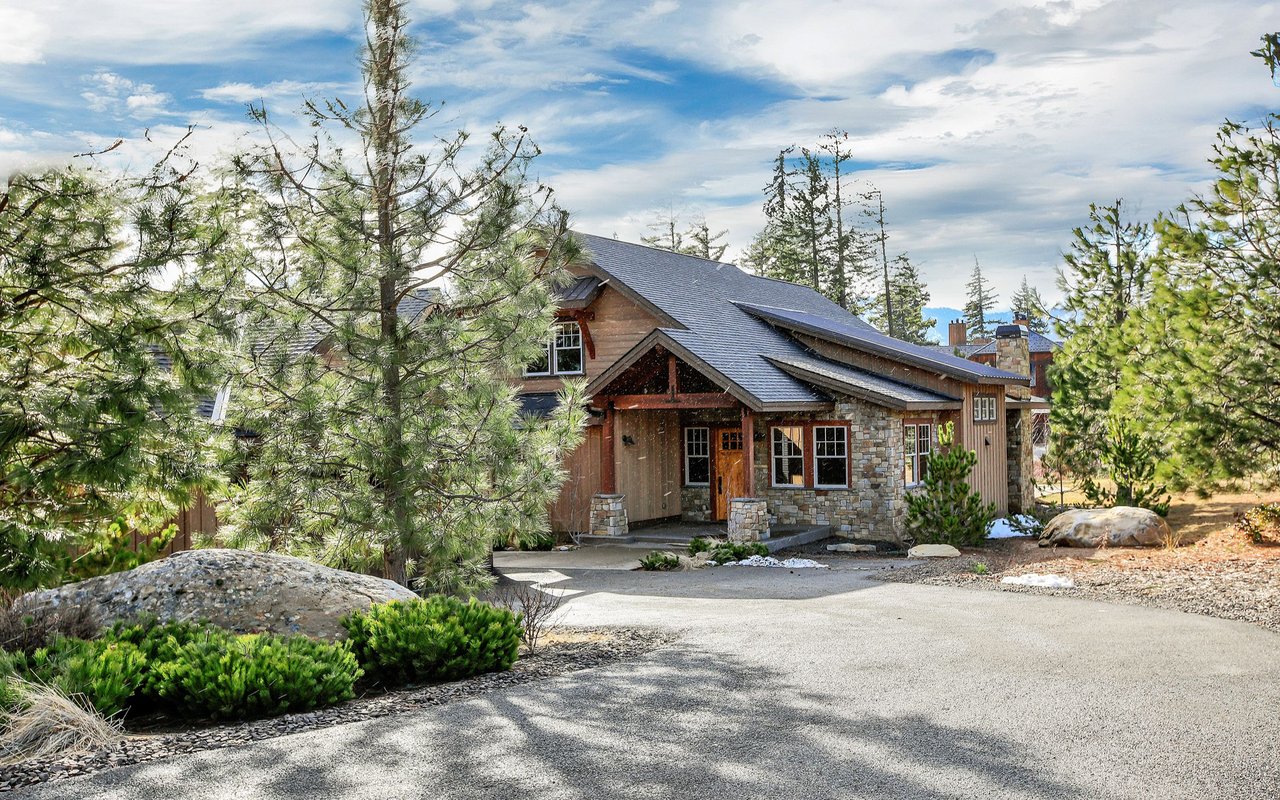 Wood-and-stone mountain home among pine trees, with a paved driveway and distant mountain ridge.