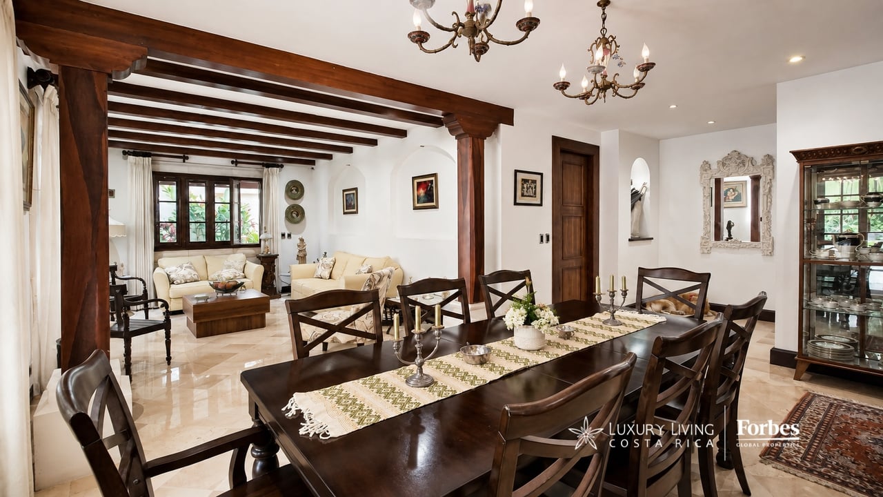 Dining and living room with a dark wood table, exposed beams, chandeliers, and cream sofas.