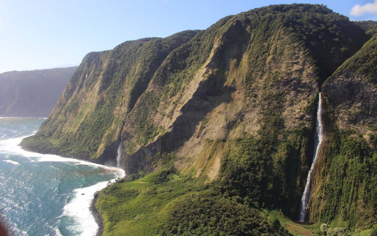 Green coastal cliffs with two waterfalls dropping toward a rocky shoreline and ocean.