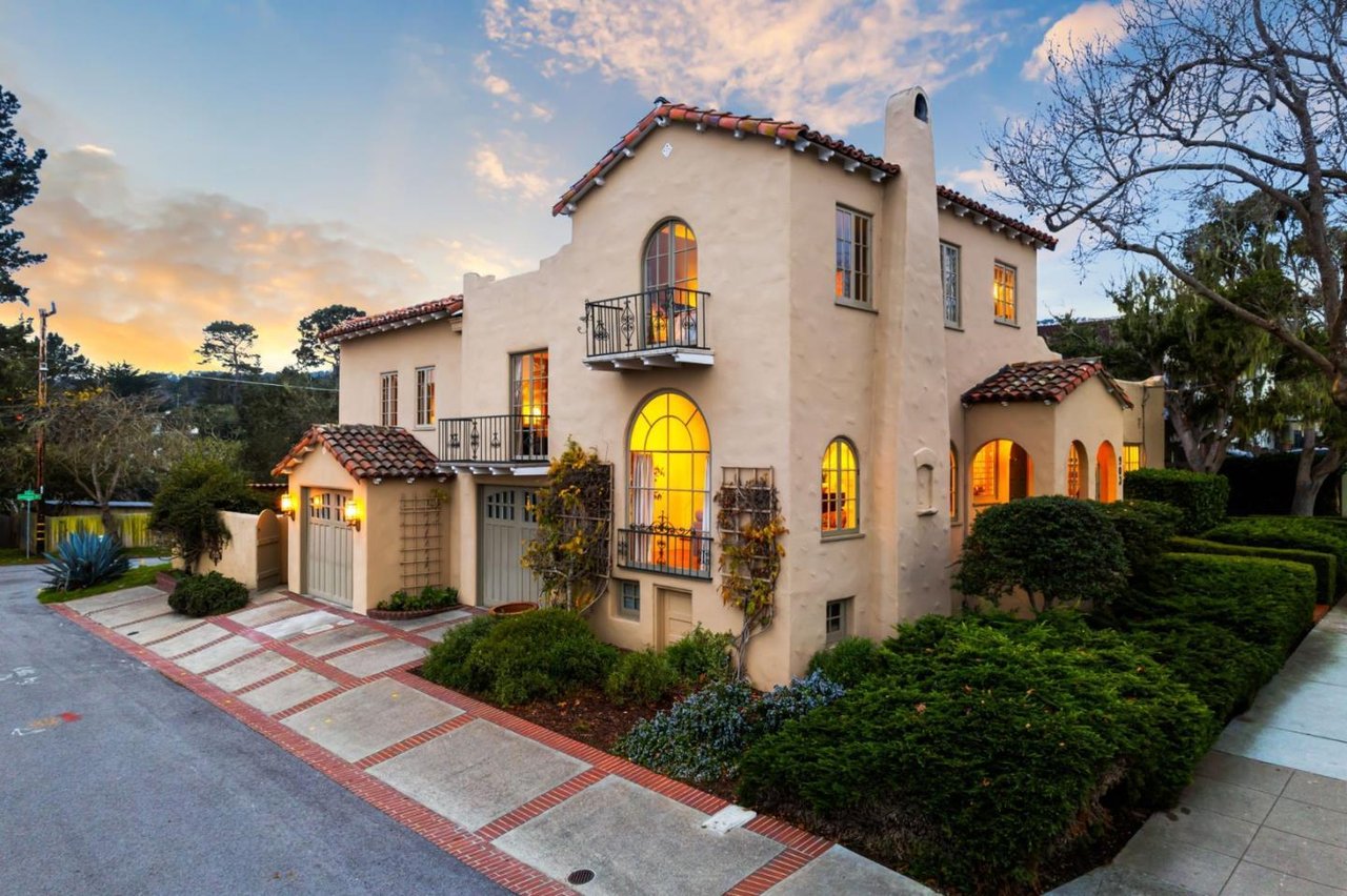 Mediterranean-style home with glowing arched windows, tile roof, balconies, and manicured hedges at dusk.