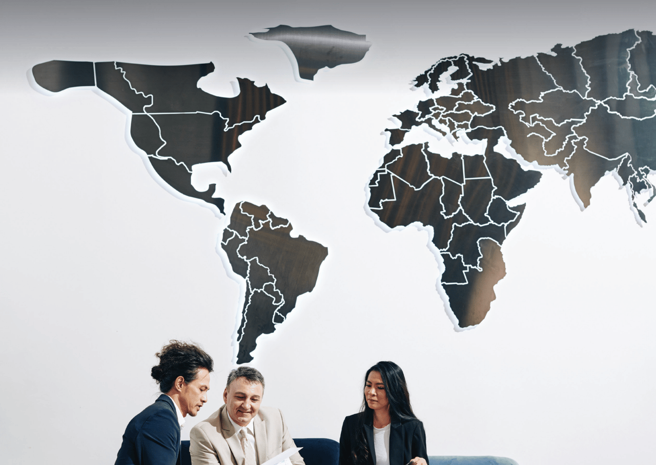 A modern world-map wall installation behind three professionals discussing global real estate strategy, representing international buyers and cross-border luxury property markets.
