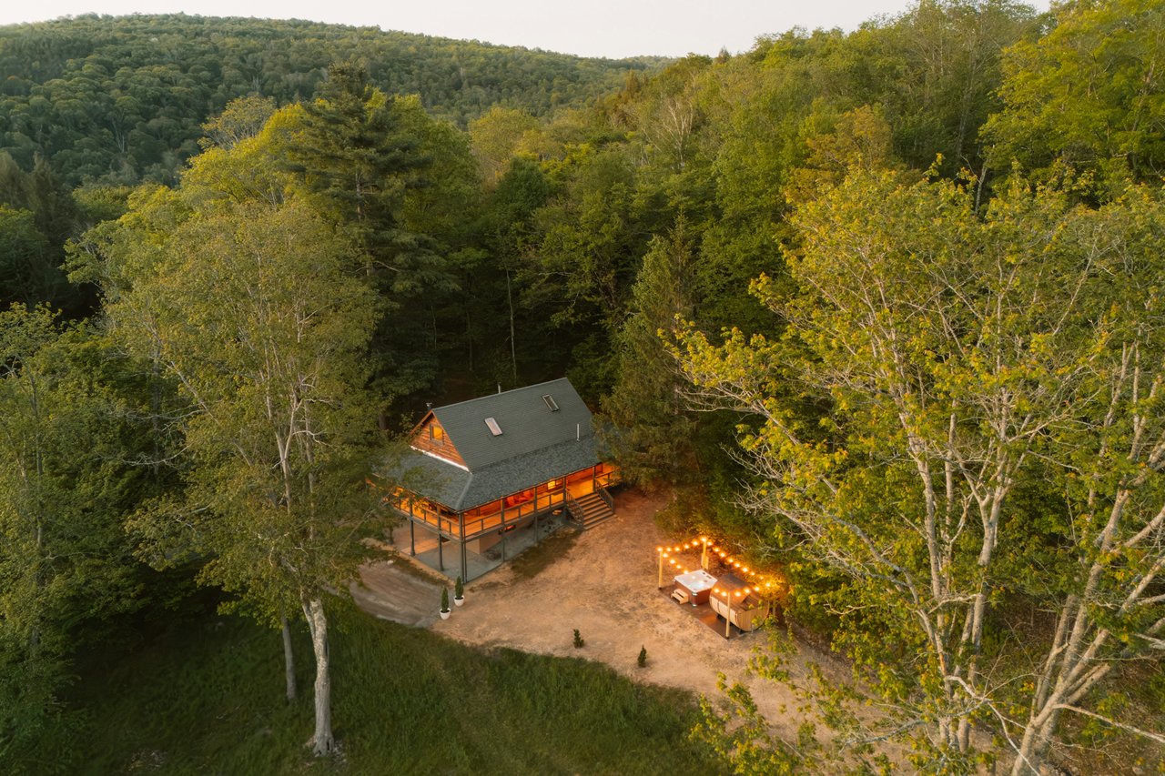 Cabin with a wraparound deck and lit outdoor seating, surrounded by autumn forest.