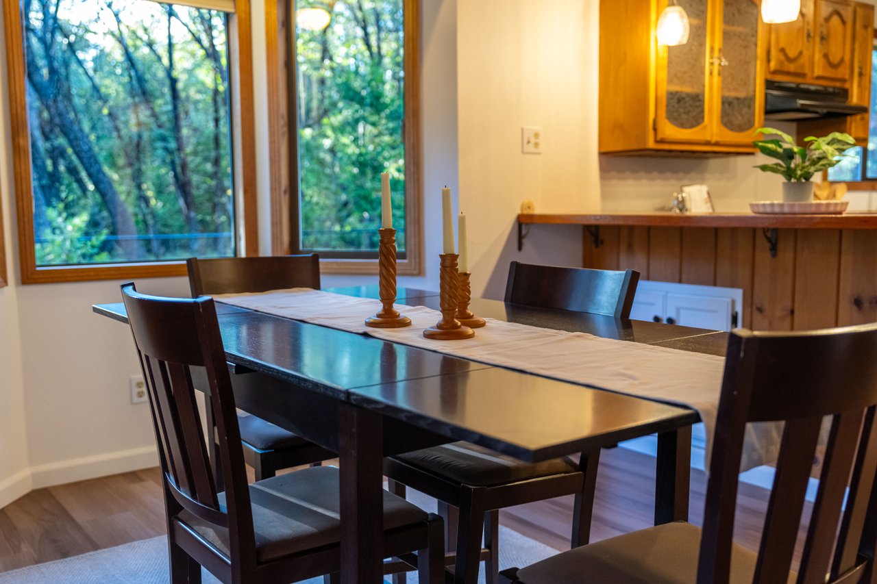 Dining area with a dark table beside large windows overlooking trees and a kitchen pass-through.