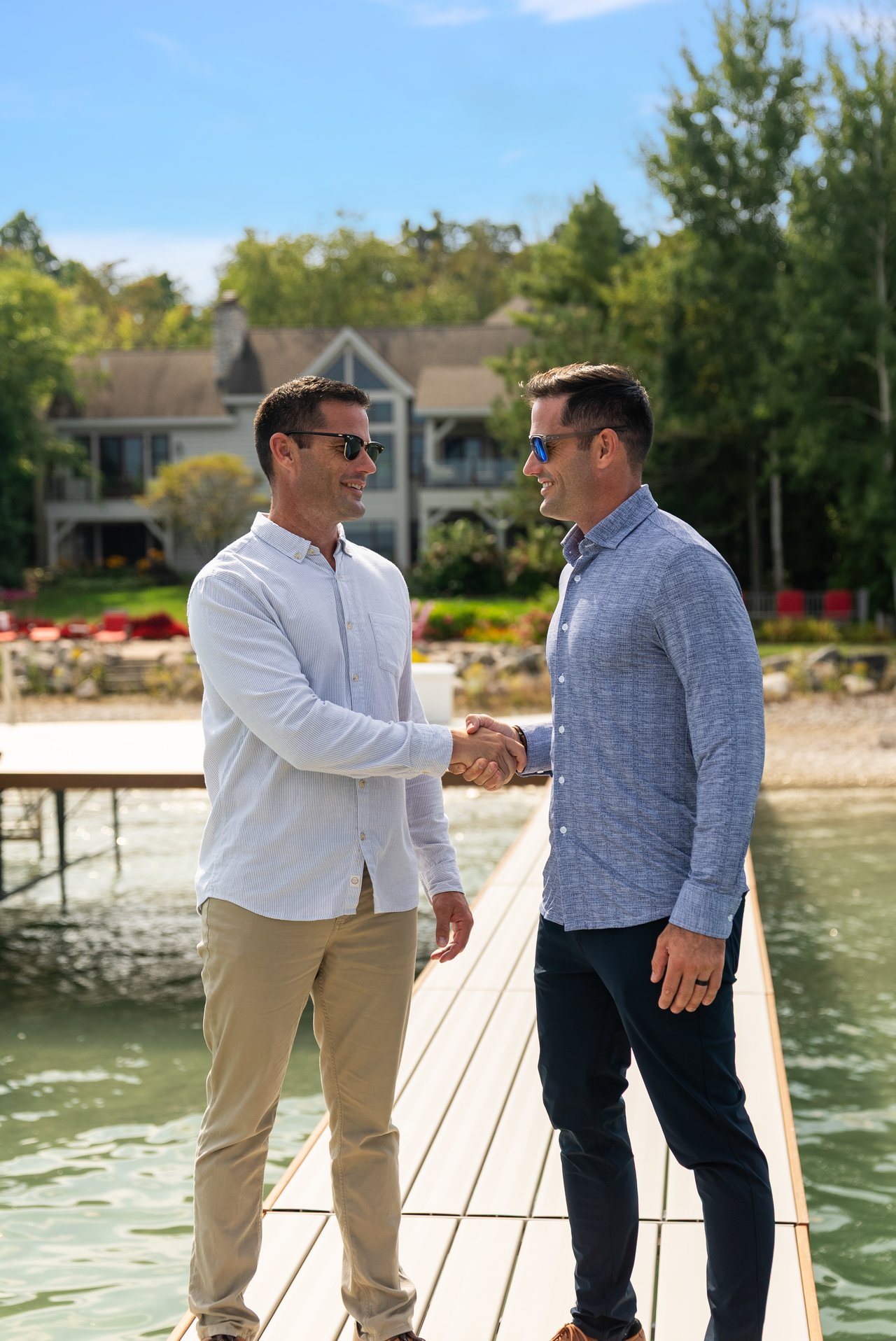 Two men shake hands on a lakeside dock in front of a large house.