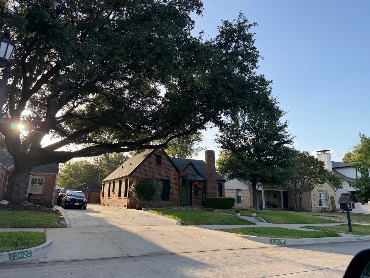 Red-brick gabled house with a tall chimney beneath a sprawling oak tree on a residential street.