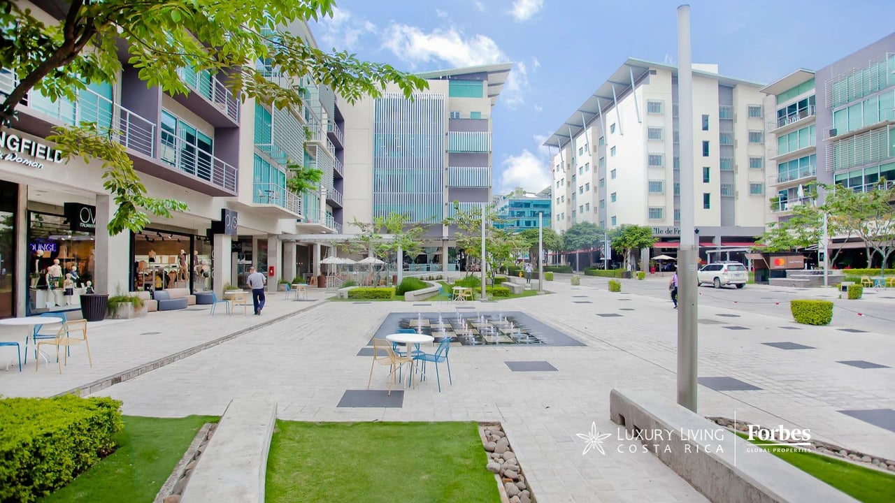 Avenida Escazú plaza with modern mixed-use buildings, storefronts, and a central fountain.