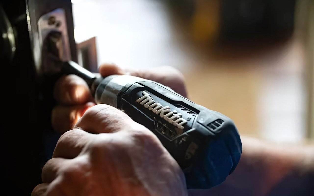  A close-up shot focuses on a pair of hands holding a black and gray Makita brand electric screwdriver. 