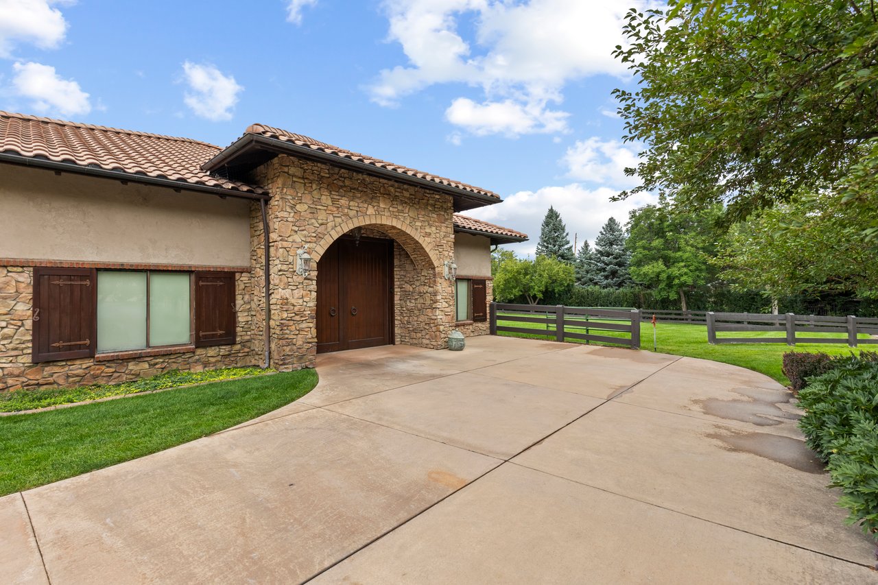 Stone barn with an arched wooden entrance and tiled roof beside a fenced lawn.