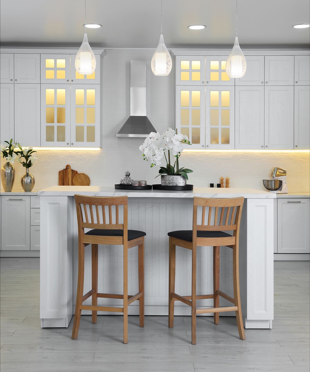 White kitchen with a large island, two wooden stools, glass-front cabinets, and pendant lights