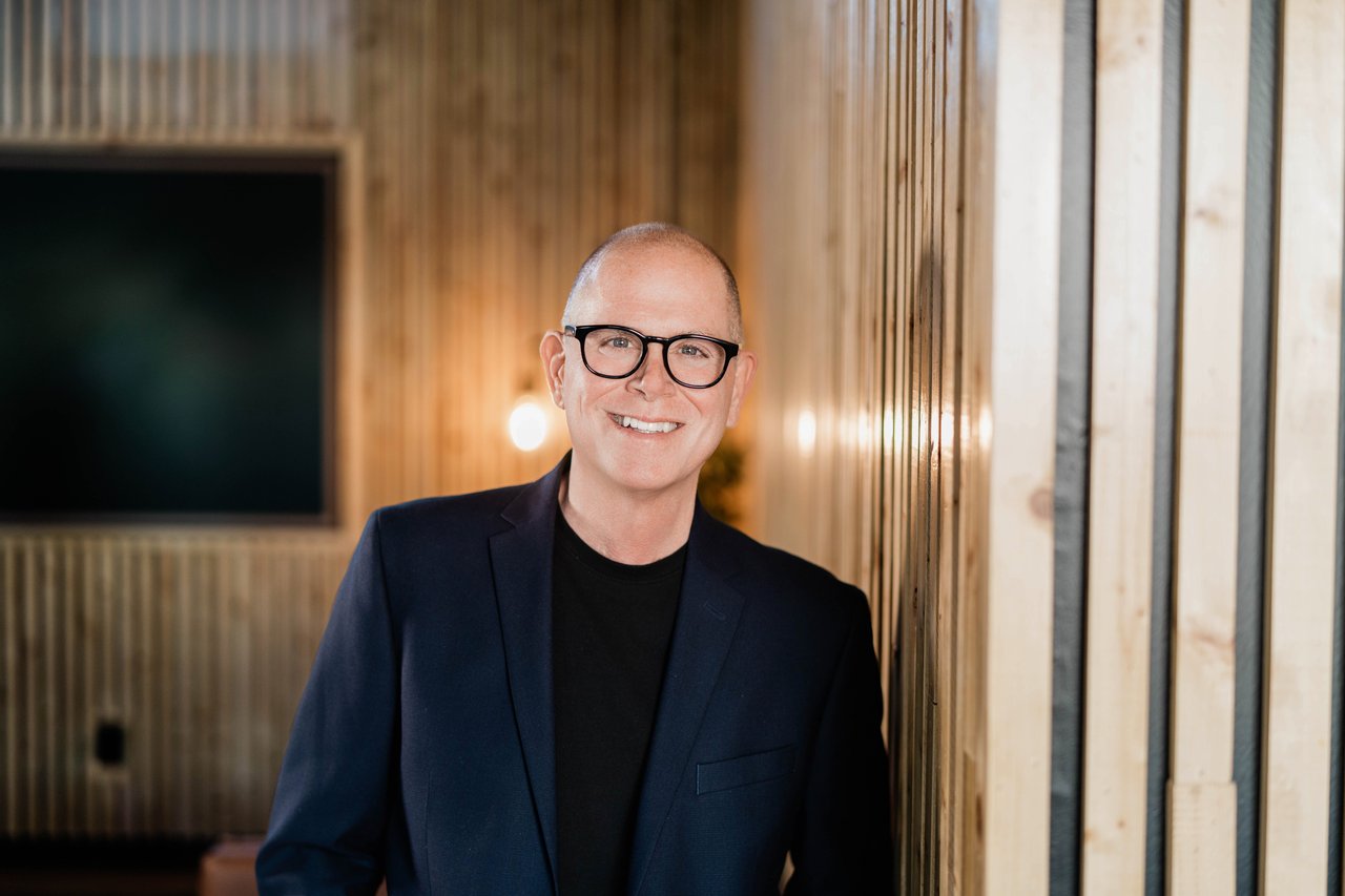Smiling man in black glasses and a navy blazer against warm wood paneling.