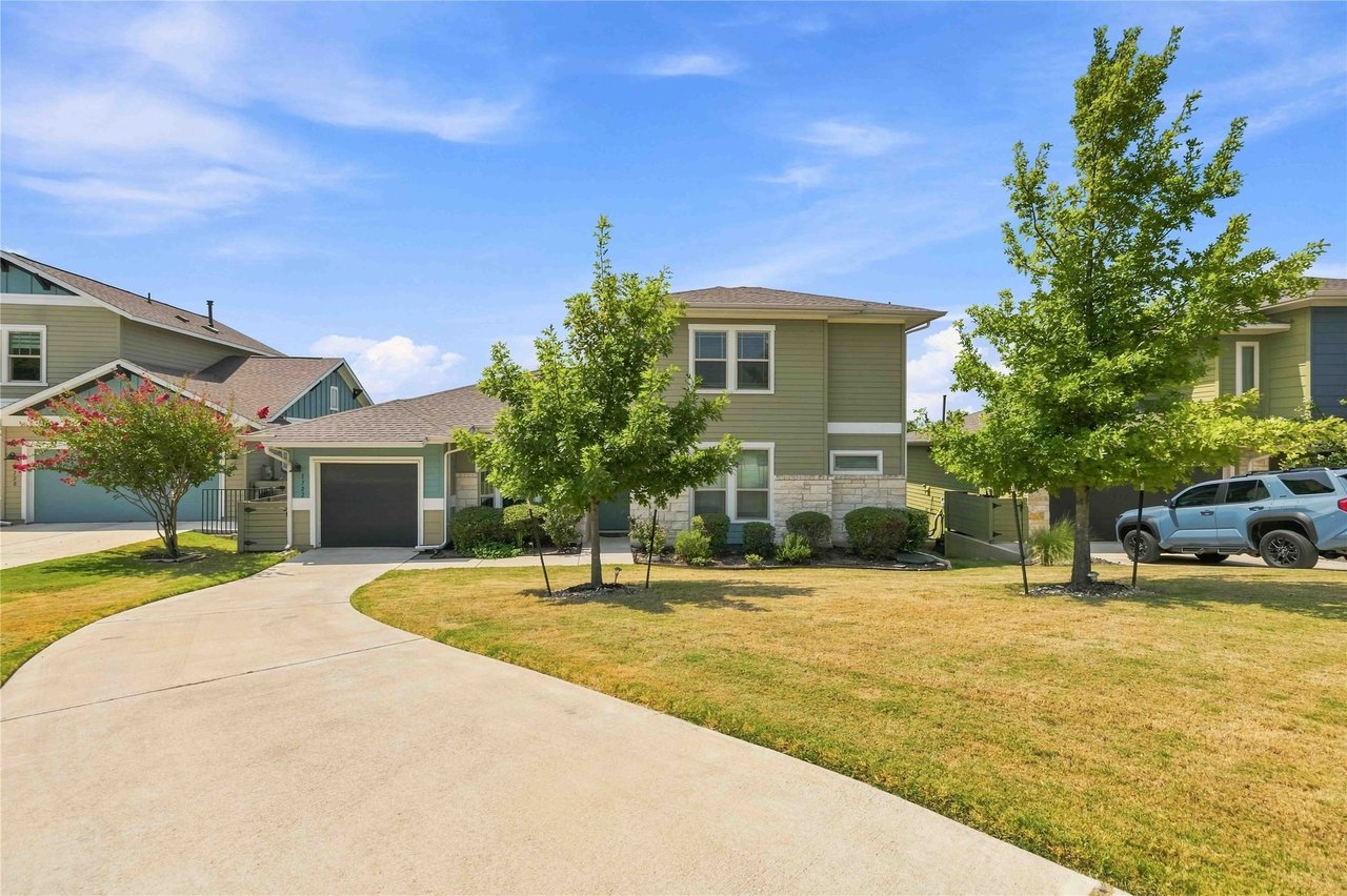 Sage-green two-story home with stone facade, attached garage, curved driveway, and mature trees.