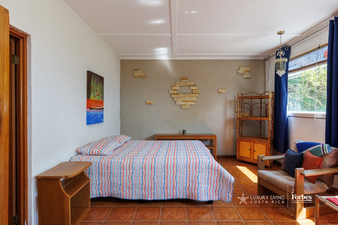 Furnished bedroom with a patterned bedspread, terracotta tile floor, and bright window with blue curtains.