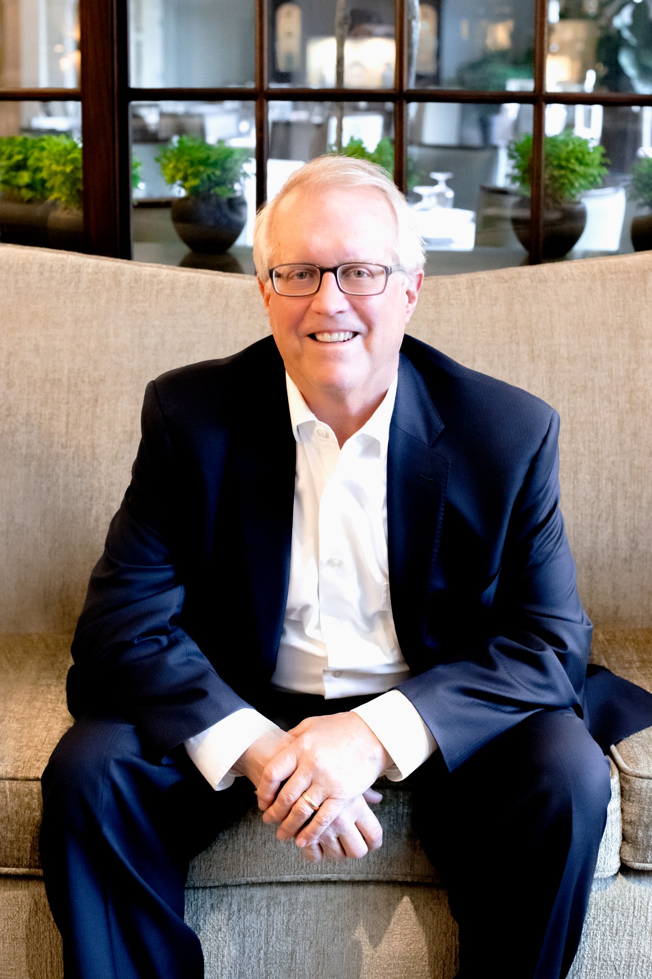Bob Seymour seated on a beige sofa in a navy suit and white shirt, smiling at the camera.