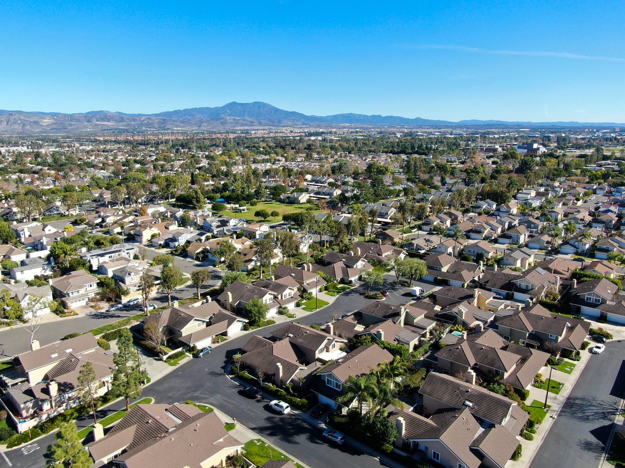 Aerial view of a suburban neighborhood with tree-lined streets, a green space, and distant mountains.